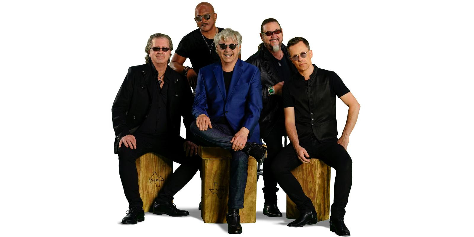 All members of the band are sat in a tight circle on wooden stools. Steve Miller is in the middle,smiling brightly, with a dark blue blazer while all other members are in all black. Everyone is in sunglasses.